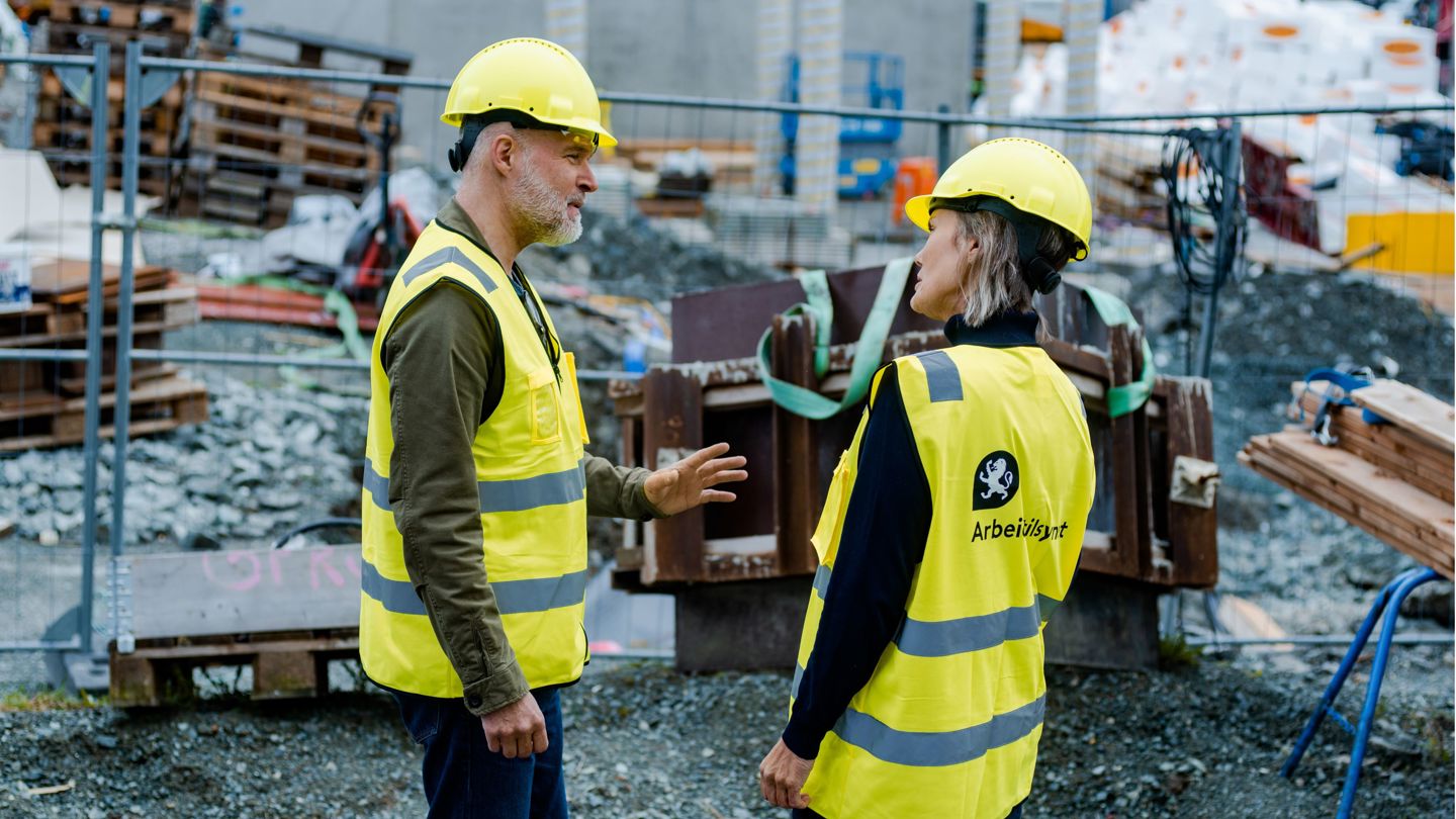 En mann og en kvinne på en byggeplass, med gul refleksvest og hjelm. Arbeidstilsynet sin logo på refleksvest.